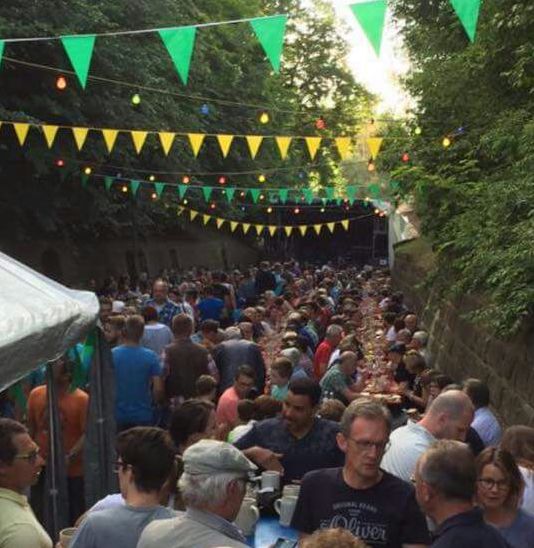 Volle Biertischreihen in der Gasse, darüber Wimpelketten und bunte Lichter